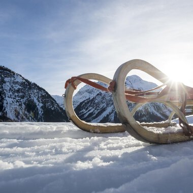 A sled lies in the snow, surrounded by a wintry mountain landscape. The sun shines out from behind the mountains. | © Kleinwalsertal Tourismus | Dominik Berchtold