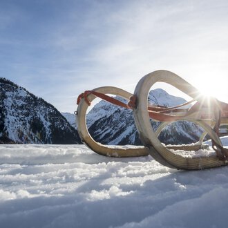 Ein Schlitten steht im Schnee, umgeben von schneebedeckten Bergen. Die Sonne strahlt im Hintergrund und erzeugt eine malerische Winterlandschaft. | © Kleinwalsertal Tourismus | Dominik Berchtold