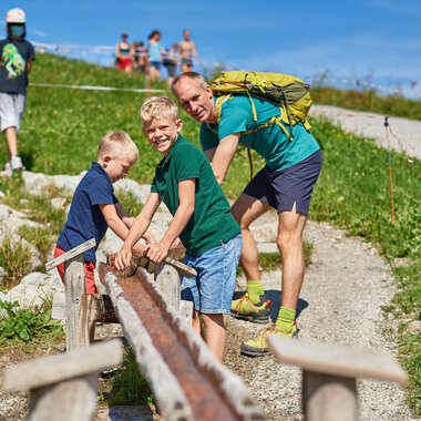Father and his two sons are playing on a path outdoors. In the background, more people can be seen, and the surroundings are green and sunny. | © OBERSTDORF · KLEINWALSERTAL BERGBAHNEN
