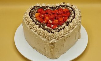 A heart-shaped cake with a creamy filling, garnished with chocolate and fresh strawberries. The background is a solid color that highlights the cake.