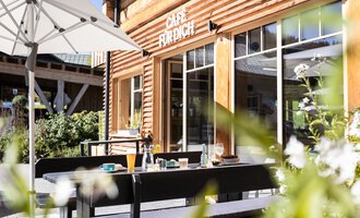 Ein einladendes Café mit Holzfassade und einem sonnigen Außenbereich. Tische sind bereit für Gäste, um die frische Luft zu genießen.