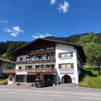 Ein charmantes Gebäude im alpinen Stil mit verschiedenen Balkonen. Im Hintergrund sind grüne Hügel und ein blauer Himmel zu sehen. | © Kleinwalsertal Tourismus | Nicole Lughammer