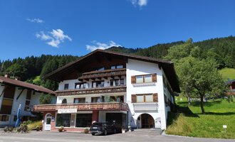 Ein charmantes Gebäude im alpinen Stil mit verschiedenen Balkonen. Im Hintergrund sind grüne Hügel und ein blauer Himmel zu sehen. | © Kleinwalsertal Tourismus | Nicole Lughammer