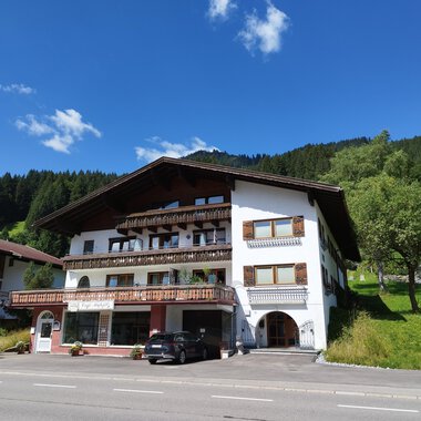Ein charmantes Gebäude im alpinen Stil mit verschiedenen Balkonen. Im Hintergrund sind grüne Hügel und ein blauer Himmel zu sehen. | © Kleinwalsertal Tourismus | Nicole Lughammer