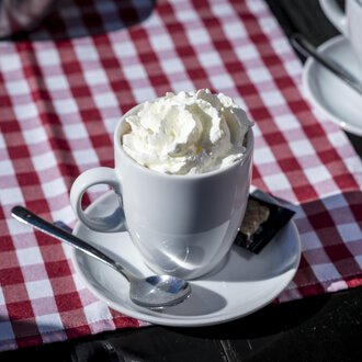 A cup with cream and a spoon is on a table with a red-and-white checkered tablecloth. In the background, more cups can be seen. | © Kleinwalsertal Tourismus | Dominik Berchtold