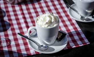A cup with cream and a spoon is on a table with a red-and-white checkered tablecloth. In the background, more cups can be seen. | © Kleinwalsertal Tourismus | Dominik Berchtold