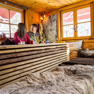 Ein gemütliches Café mit Holzmöbeln und weichen Decken. Zwei Personen sitzen am Fenster und genießen ihre Zeit miteinander. | © Kleinwalsertal Tourismus | Dominik Berchtold