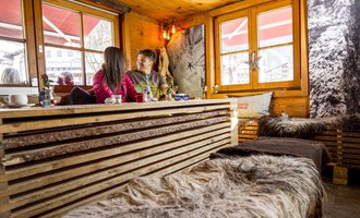 Ein gemütliches Café mit Holzmöbeln und weichen Decken. Zwei Personen sitzen am Fenster und genießen ihre Zeit miteinander. | © Kleinwalsertal Tourismus | Dominik Berchtold