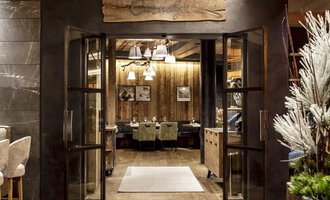 An inviting entrance to a restaurant with wooden details and warm lighting. The atmosphere is cozy and welcoming. | © Travel Charme Ifen Hotel