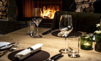 An elegant table with dishes and glasses, decorated for a dinner. In the background, a fireplace is burning, creating a cozy atmosphere. | © Travel Charme Ifen Hotel