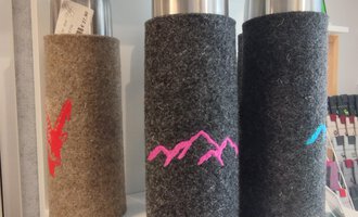 Three thermos bottles with felt covers in different colors are standing on a shelf. The bottles have colored mountain motifs on the covers. | © Chrömle Schönes aus Filz | N. Lughammer