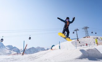 Ein Snowboarder springt über einen Schneehügel vor einer klaren Berglandschaft. Im Hintergrund sind Seilbahnen und weitere Skifahrer zu sehen. | © OBERSTDORF · KLEINWALSERTAL BERGBAHNEN