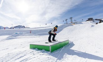 Ein Snowboarder springt über eine grüne Box im Schnee. Im Hintergrund sind Berge und Skilifte sichtbar. | © OBERSTDORF · KLEINWALSERTAL BERGBAHNEN
