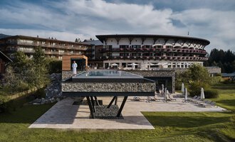 A modern hotel with a large glass pool is in the foreground. The surrounding landscape is green and inviting. | © Travel Charme Ifen Hotel