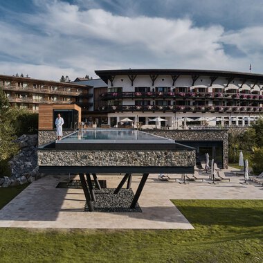 A modern hotel with a large glass pool is in the foreground. The surrounding landscape is green and inviting. | © Travel Charme Ifen Hotel