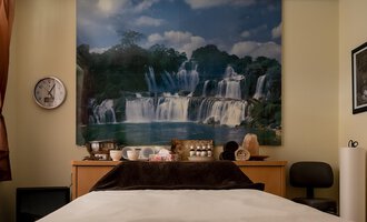 A cozy room with a bed and a large wall painting of waterfalls. On the table, there are various decorative items and a clock visible. | © IFA Hotel Alpenhof Wildental | Andreas Froschauer