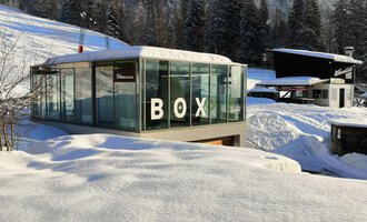 A modern glass building with the inscription "BOX" stands amidst a snow-covered landscape. In the background, snow-covered trees and additional buildings can be seen. | © Sport Kessler GmbH