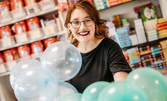 A young woman with glasses smiles and holds colorful balloons in a store. In the background, bookshelves and other items are visible.