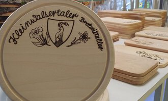 A wooden board with the print "Kleinwalsertaler Brotmitteller" is visible in the foreground. In the background, additional wooden boards are lying on a table. | © Der Bunte Laden | Romana Volderauer