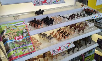 A selection of different toy horses in various colors and sizes is displayed on a shelf. Below are also toy cards and catalogs for further illustration. | © Der Bunte Laden | Romana Volderauer