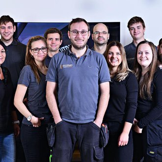 A group of twelve people stands together and smiles. They are wearing similar gray work clothing and are in a bright room. | © Der Elektriker Markus Leitgeb GmbH
