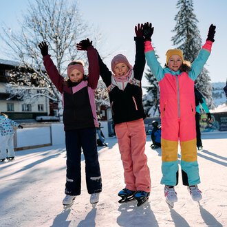 A group of cheerful children stands on an ice rink, raising their hands in the air. They are wearing colorful winter clothing and enjoying the sunny winter day. | © Kleinwalsertal Tourismus | Oliver Farys