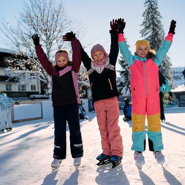A group of cheerful children stands on an ice rink, raising their hands in the air. They are wearing colorful winter clothing and enjoying the sunny winter day. | © Kleinwalsertal Tourismus | Oliver Farys