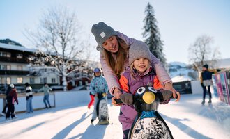 A mother and her child are riding on a penguin ice surface. In the background, winter landscapes and people skating can be seen. | © Kleinwalsertal Tourismus | Oliver Farys