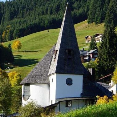 A picturesque church with a tall spire stands in a green landscape. In the background, there are gentle hills and some houses. | © Ev. Kirche Kleinwalsertal