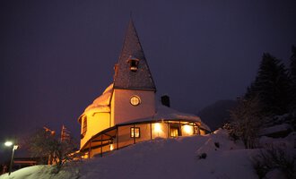 A charming snow-covered house with a peaked roof in a wintry landscape. The warm lights in the building create a cozy atmosphere in the darkness. | © Ev. Kirche Kleinwalsertal