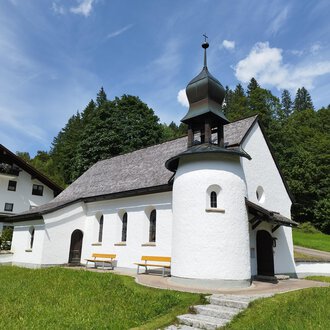 Eine kleine, weiße Kapelle mit einem schwarzen Dach inmitten von Grün. Im Hintergrund sind Bäume und eine Ferienunterkunft zu sehen. | © Kleinwalsertal Tourismus | N. Lughammer