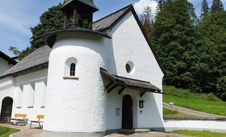 Eine kleine, weiße Kirche mit einem markanten Dach und einem Glockenturm. Sie befindet sich in einer grünen Umgebung mit Bäumen und klarem Himmel. | © Kleinwalsertal Tourismus | N. Lughammer