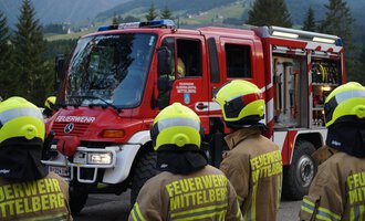Ein Feuerwehrauto steht auf einer Straße, umgeben von Feuerwehrleuten in uniformen Gelben Helmen. Die Landschaft im Hintergrund zeigt Bäume und Berge. | © Feuerwehr Mittelberg | Christoph Hilbrand