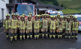 Eine Gruppe von Feuerwehrleuten in Einsatzkleidung steht vor einem Feuerwehrauto. Im Hintergrund sind ein Gebäude und eine grüne Hügelkulisse zu sehen. | © Feuerwehr Mittelberg | Christoph Hilbrand
