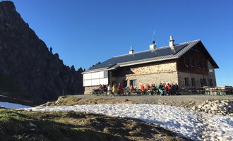 Eine Berghütte in den Alpen umgeben von schneebedecktem Boden. Viele Menschen sitzen draußen und genießen die Sonne unter blauen Himmel. | © Kleinwalsertal Tourismus