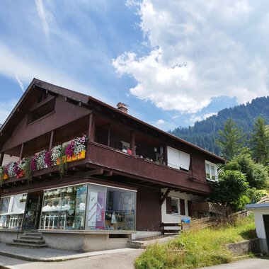A charming house with a blooming balcony in the mountains. The surroundings are green, and the cloudy sky creates a beautiful atmosphere. | © Kleinwalsertal Tourismus | N. Lughammer