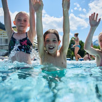 Drei fröhliche Kinder planschen im Schwimmbecken und haben viel Spaß. Sie heben die Hände in die Luft und genießen die Sonne. | © Kleinwalsertal Tourismus | Oliver Farys