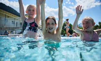 Drei fröhliche Kinder planschen im Schwimmbecken und haben viel Spaß. Sie heben die Hände in die Luft und genießen die Sonne. | © Kleinwalsertal Tourismus | Oliver Farys
