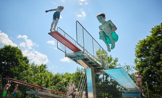 Kinder springen von einem Sprungturm ins Wasser. Es ist ein sonniger Tag mit blauem Himmel und grünen Bäumen im Hintergrund. | © Kleinwalsertal Tourismus | Oliver Farys