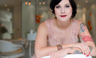 A woman with tattoos is sitting smiling in a modern room. She is wearing an elegant pink dress and has a chic look. | © Fräulein Sugar Coiffeur | Sandra Bromm