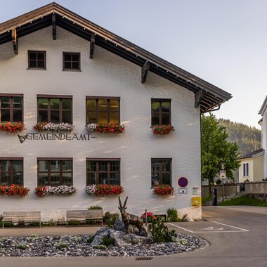 A charming community building with blooming flower boxes. In the background, additional buildings and trees can be seen. | © Kleinwalsertal Tourismus |  Steffen Berschin