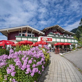 A cozy restaurant with a red facade and blooming flowers in the foreground. Red umbrellas provide shade for the guests. | © Gasthof Alpenblick | Theodor Pinn