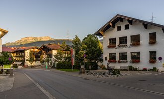 A picturesque street view of a small village in the Alps, surrounded by mountains. The charming houses are adorned with flowers, creating an inviting atmosphere. | © Kleinwalsertal Tourismus |  Steffen Berschin
