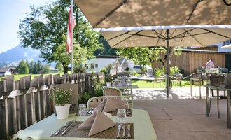 An inviting terrace with covered tables and sunshades. In the background, green trees and a beautiful landscape can be seen.
