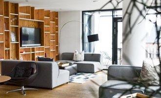 A modern living room with gray sofas and a wooden shelf. Large windows provide plenty of natural light.