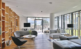 A modern living room with large windows and plenty of natural light. The furniture is stylish and minimalist, featuring a wooden bookshelf and comfortable seating areas.