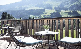 A terrace with modern chairs and tables. In the background, green hills and mountains can be seen.