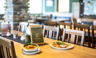 A cozy restaurant with wooden chairs and a clean table. On the table lies a menu, next to it is a plate with meat and vegetables as well as a bowl of rice. | © OBERSTDORF·KLEINWALSERTAL BERGBAHNEN