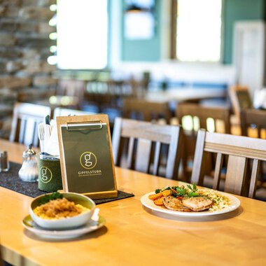 Ein gemütliches Restaurant mit Holzstühlen und einem sauberen Tisch. Auf dem Tisch liegt eine Speisekarte, daneben befinden sich ein Teller mit Fleisch und Gemüse sowie eine Schüssel mit Reis. | © OBERSTDORF·KLEINWALSERTAL BERGBAHNEN