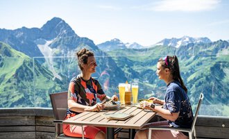Two women are sitting at a table enjoying drinks and food. In the background, green mountains and a blue sky are visible. | © Oberstdorf · KLEINWALSERTAL BERGBAHNEN